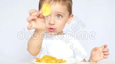 一个喜欢吃薯片的孩子坐在白色背景的桌子旁。 男孩正在吃薯片，微笑着。